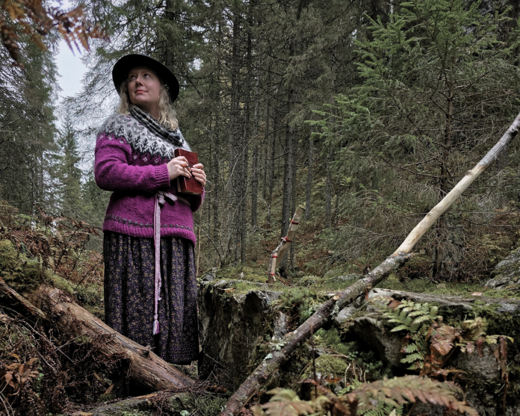 ninni-witches-north-gorge woman in sweater and skirt staring next to a flat boulder in forest